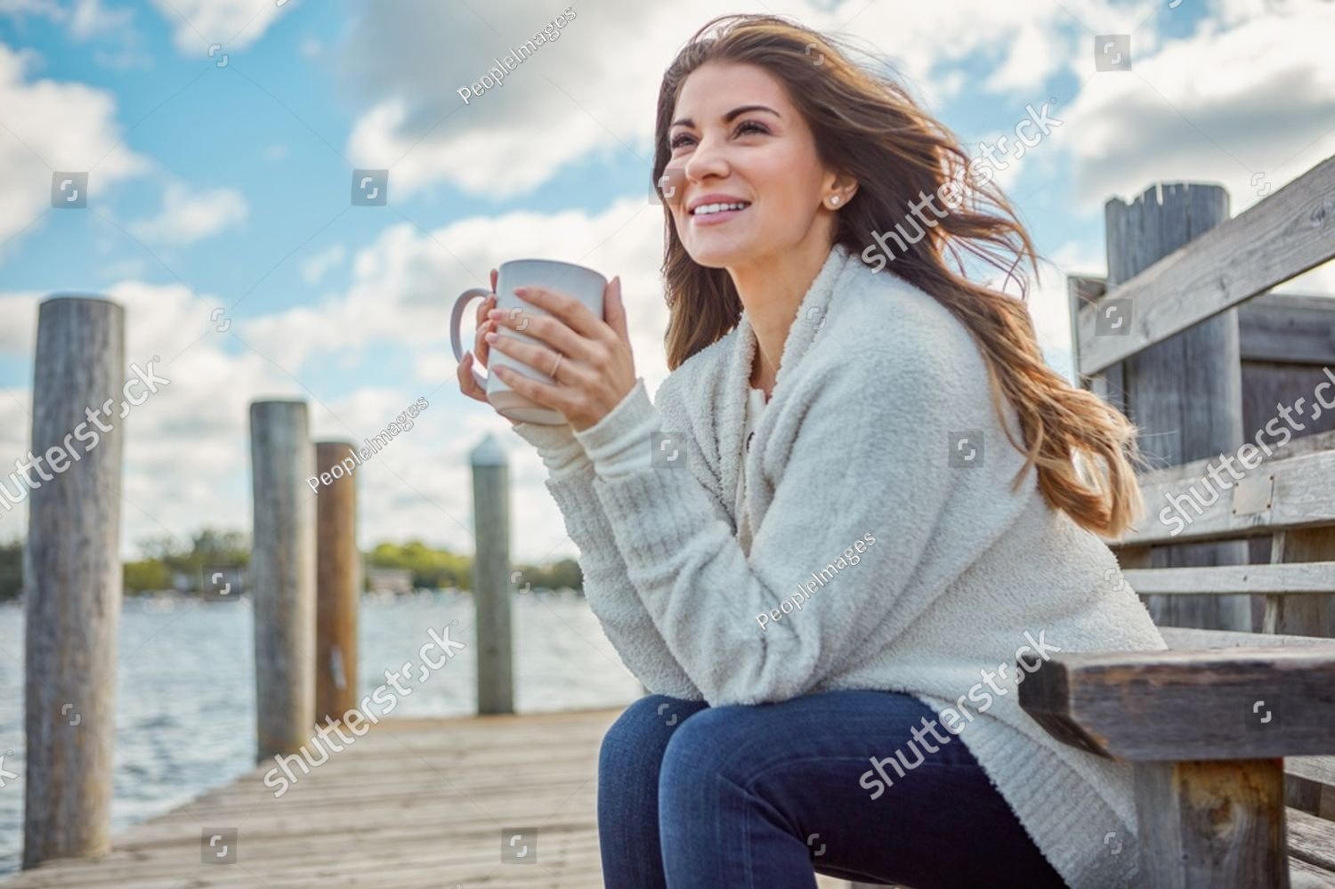 A woman on a dock holding a warm mug, looking out over the water